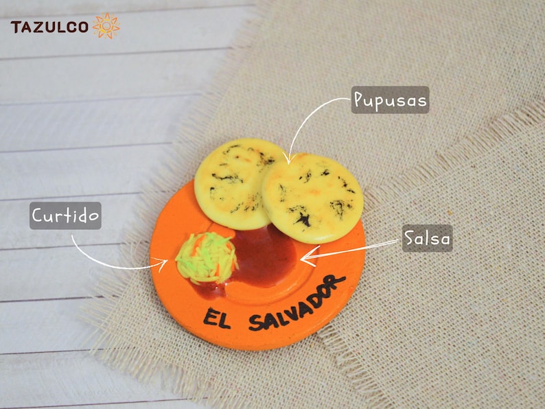 Puede incluir: Primer plano de un peque&ntilde;o plato naranja con las palabras "El Salvador" escritas en &eacute;l. El plato tiene dos pupusas amarillas, un poco de salsa roja y un poco de curtido verde. El plato est&aacute; sobre un fondo de arpillera beige.