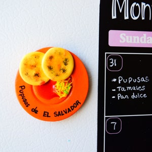 Puede incluir: Un plato de cer&aacute;mica naranja con dos pupusas amarillas y salsa roja. El plato tiene el texto "Pupusas de El Salvador" escrito en &eacute;l.
