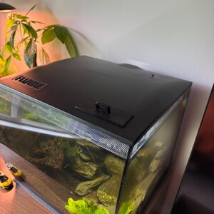 May include: A black Fluval aquarium with a curved glass front. The tank contains rocks, plants, and water. The top of the aquarium is black with a small access door. The aquarium sits on a wooden surface.