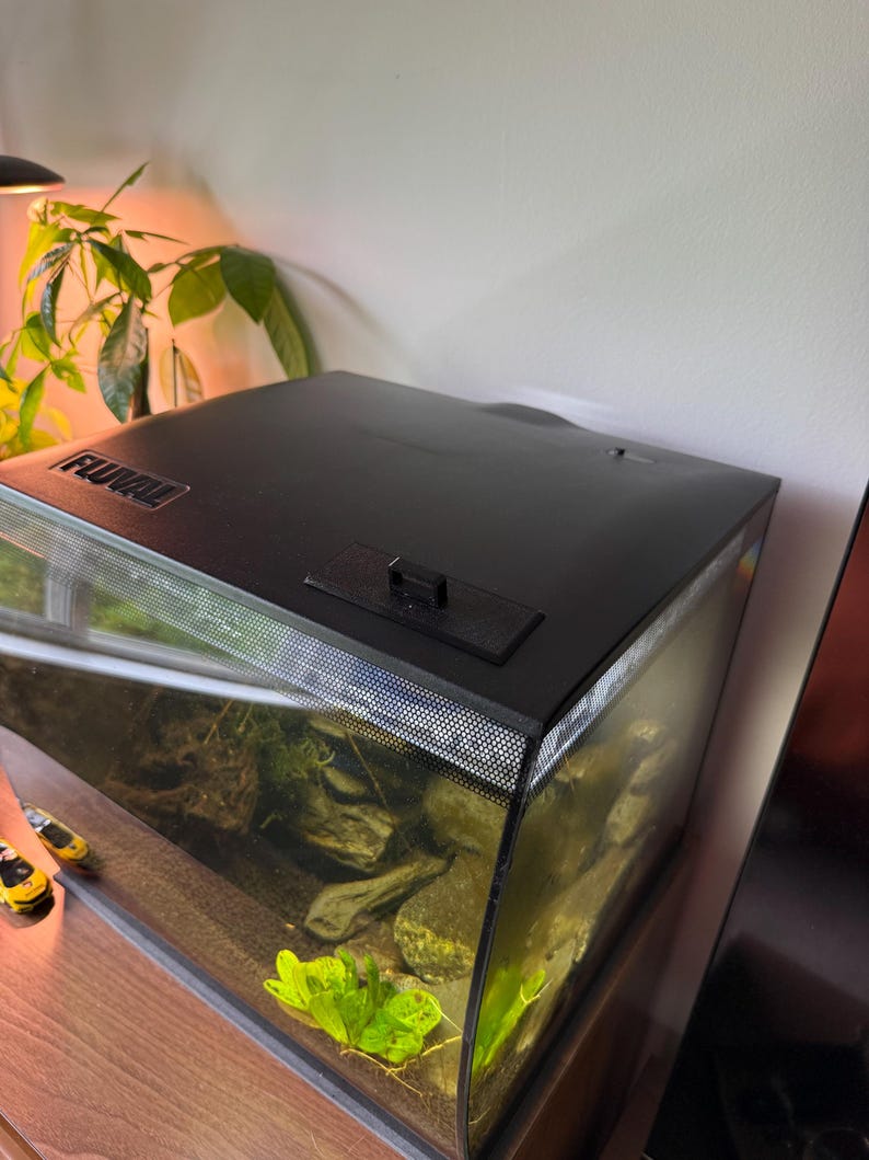 May include: A black Fluval aquarium with a curved glass front. The tank contains rocks, plants, and water. The top of the aquarium is black with a small access door. The aquarium sits on a wooden surface.