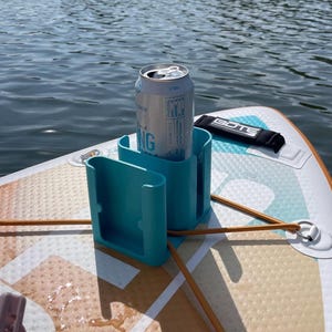 Puede incluir: Un portavasos turquesa con una lata de agua con gas, montado en una tabla de paddle surf. La tabla tiene un diseño colorido, con una correa negra y la palabra "BOTE" visible.