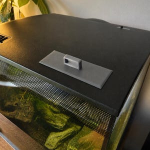 May include: A black aquarium with a mesh screen and a gray handle on top. The tank contains rocks and aquatic plants. The top of the aquarium is black and has a textured surface.