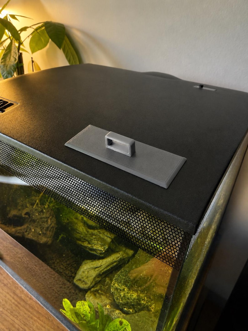 May include: A black aquarium with a mesh screen and a gray handle on top. The tank contains rocks and aquatic plants. The top of the aquarium is black and has a textured surface.