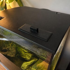 May include: A black aquarium tank with a mesh screen and a black lid. The tank contains rocks and aquatic plants. The lid has a handle and a rectangular platform. The tank is placed on a wooden surface.