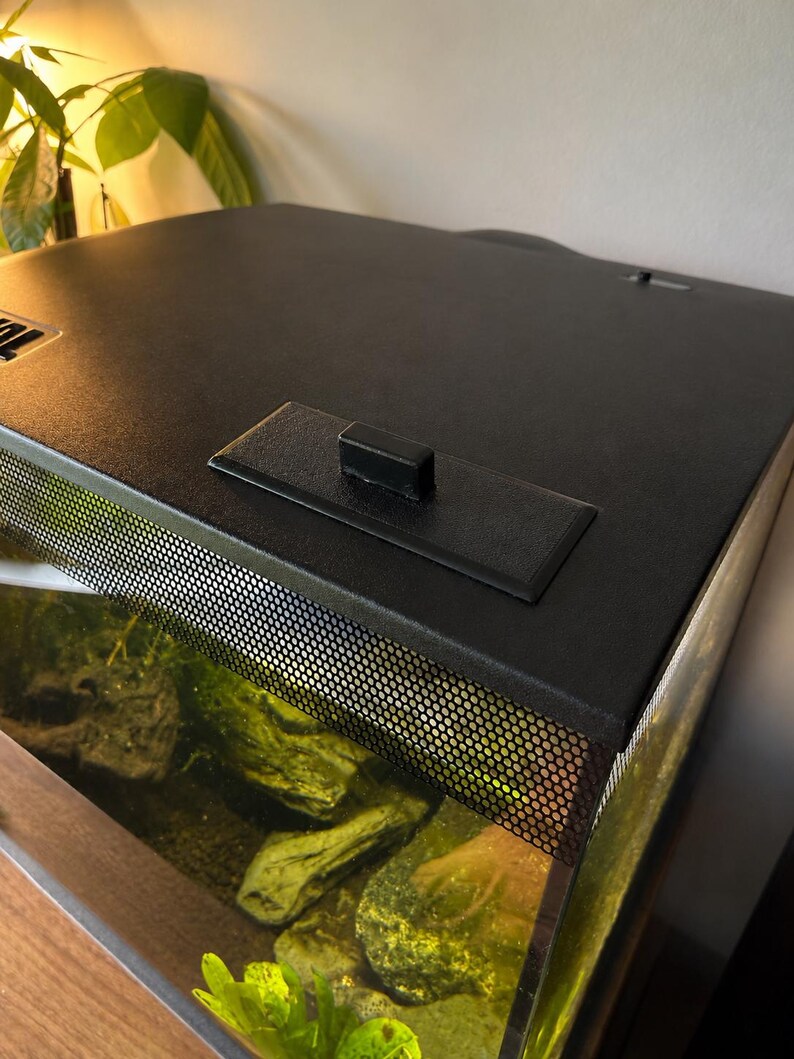 May include: A black aquarium tank with a mesh screen and a black lid. The tank contains rocks and aquatic plants. The lid has a handle and a rectangular platform. The tank is placed on a wooden surface.