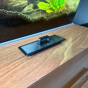 May include: A black rectangular aquarium lid with a handle sits on a wooden surface. The lid is in front of a glass aquarium with green plants and rocks inside. The wooden surface has a natural wood grain.