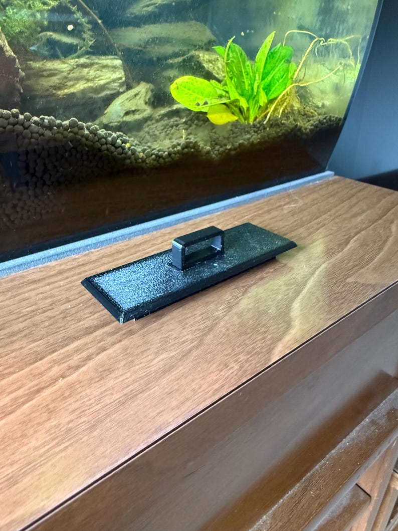 May include: A black rectangular aquarium lid with a handle sits on a wooden surface. The lid is in front of a glass aquarium with green plants and rocks inside. The wooden surface has a natural wood grain.