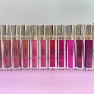 May include: A row of 12 clear tubes with gold lids containing different shades of lipstick. The tubes are labeled "Lipstick Organic" and are arranged on a pink surface.