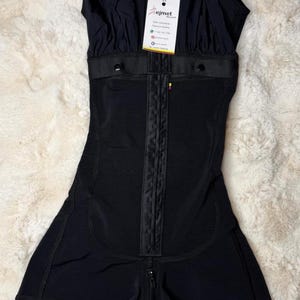 May include: A black compression bodysuit featuring a front hook-and-eye closure, wide shoulder straps, and lace trim at the leg openings. A white product tag with the text Bejmet is attached to the neckline, displaying contact information.