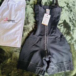 May include: A black compression bodysuit with adjustable shoulder straps, a front hook-and-eye closure, and a zipper fly. The garment features a textured lace-like hem and includes a white drawstring storage bag with the logo Bejmet printed on it.