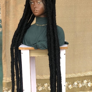 May include: A black wig featuring long, thick, textured faux locs styled with a center part. The wig is displayed on a mannequin head wearing a dark green mock neck top and a thin gold chain necklace, showcasing the length and volume of the hair.
