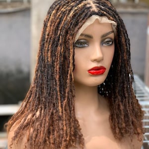 以下が含まれることがあります： ブロンドのハイライトが入った、茶色と黒のドレッドヘアのウィッグをつけたマネキンの頭。ウィッグはレースフロントで、肩までの長さのドレッドヘアにスタイリングされています。マネキンは赤い口紅を塗っており、背景はぼやけています。