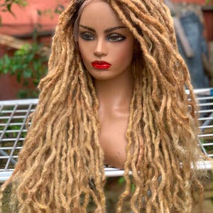 May include: A lace front wig featuring long, textured dreadlocks in a blend of blonde and light brown tones. The hair is styled with a side part, and the dreadlocks fall past the shoulders in a layered, voluminous arrangement with dark roots.