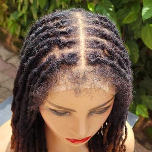 May include: A mannequin head with a dark brown and auburn dreadlock wig. The wig features a lace front and is styled with individual locs. The background includes green foliage and a red flower.
