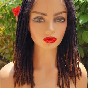 May include: A mannequin head with a dark brown and light brown dreadlock wig. The mannequin has red lipstick and is set against a backdrop of green foliage.