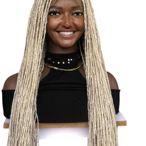 May include: A blonde dreadlock wig on a mannequin head. The wig features long, textured dreadlocks and a lace front. The mannequin is wearing a black off-the-shoulder top and a gold chain necklace. The background is white.