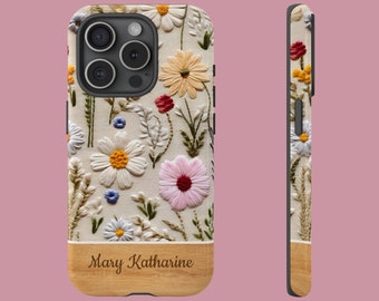 May include: A smartphone case featuring an embroidered floral pattern with white, yellow, pink, and red flowers on a cream background. The bottom section has a light wood grain texture with the name Mary Katharine printed in a dark brown script.