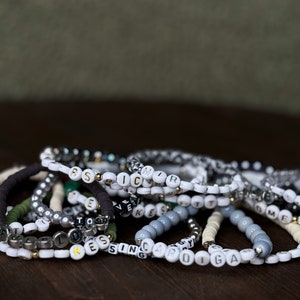 May include: A collection of beaded bracelets with white, gray, black, and gold beads. Some bracelets feature letter beads spelling out names or words. The bracelets are arranged on a dark brown wooden surface.