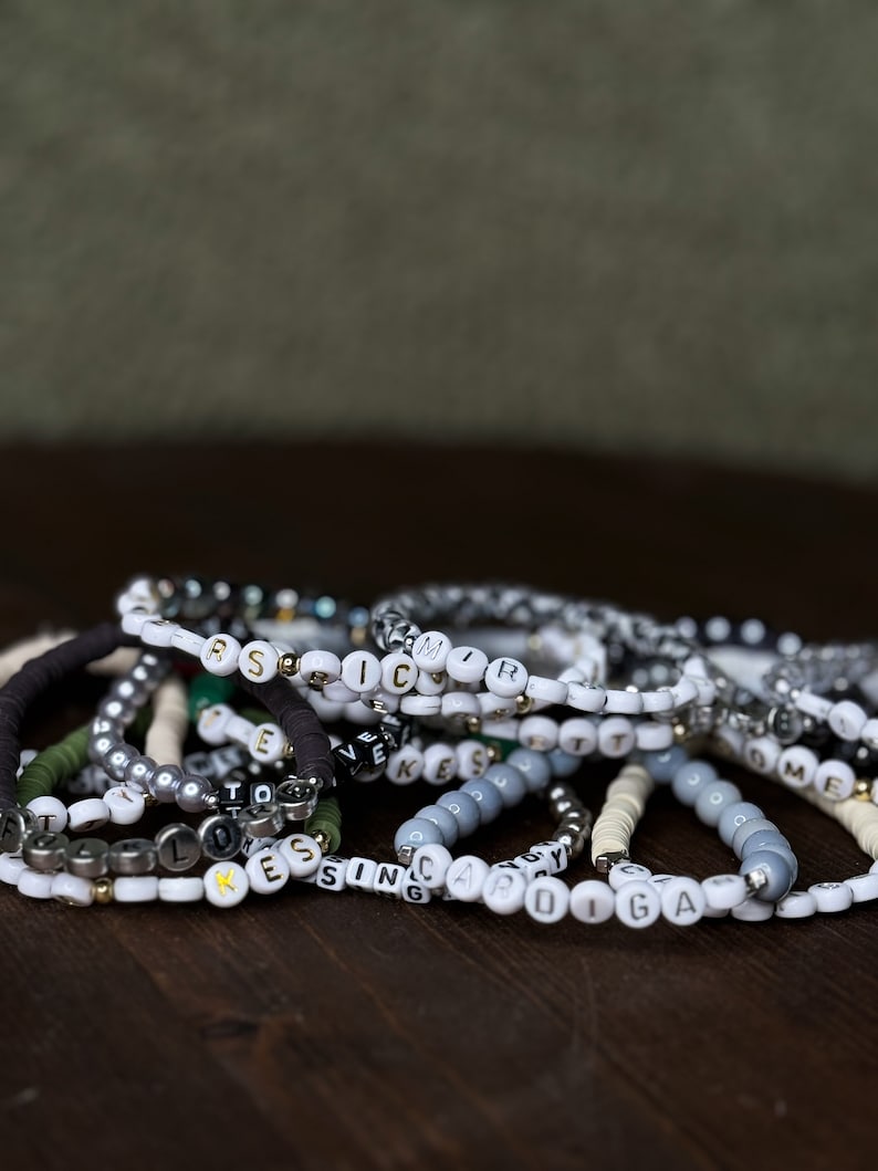 May include: A collection of beaded bracelets with white, gray, black, and gold beads. Some bracelets feature letter beads spelling out names or words. The bracelets are arranged on a dark brown wooden surface.