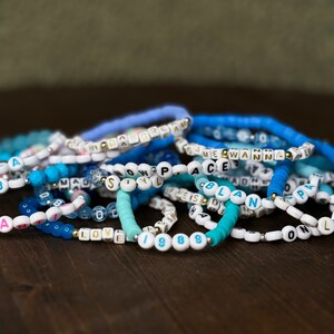 May include: A collection of beaded bracelets in shades of blue and white, with some featuring letter beads spelling out words like "LOVE", "PEACE", and "1988". The bracelets are stacked on a dark wooden surface.