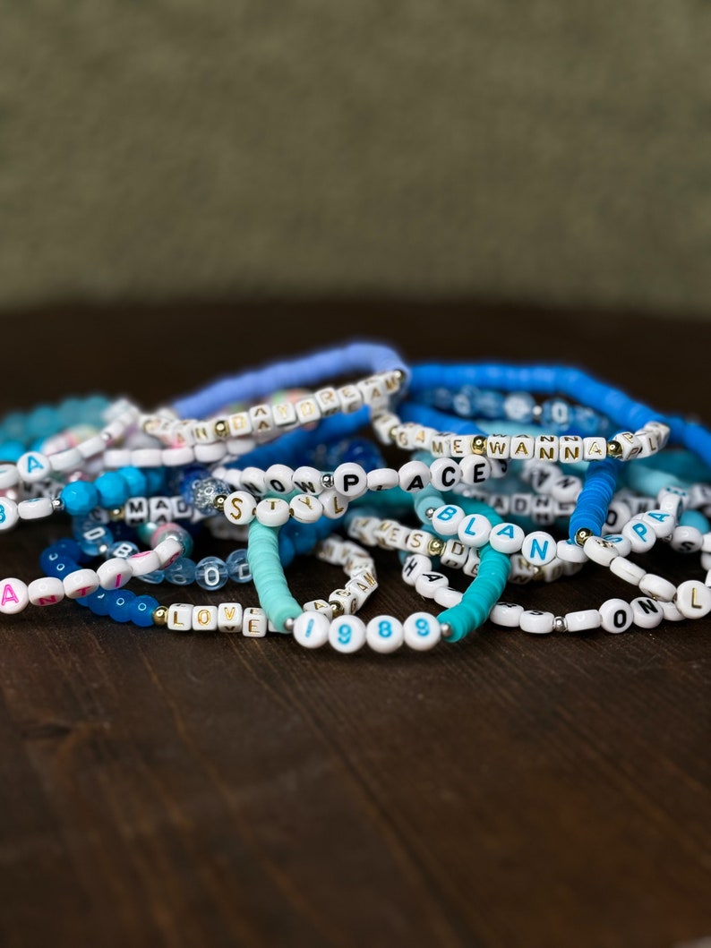 May include: A collection of beaded bracelets in shades of blue and white, with some featuring letter beads spelling out words like "LOVE", "PEACE", and "1988". The bracelets are stacked on a dark wooden surface.