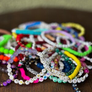 May include: A collection of colorful beaded bracelets with white letter beads spelling out words. The bracelets are in various colors, including pink, yellow, green, and purple. Some bracelets have gold accents and are arranged on a dark wooden surface.