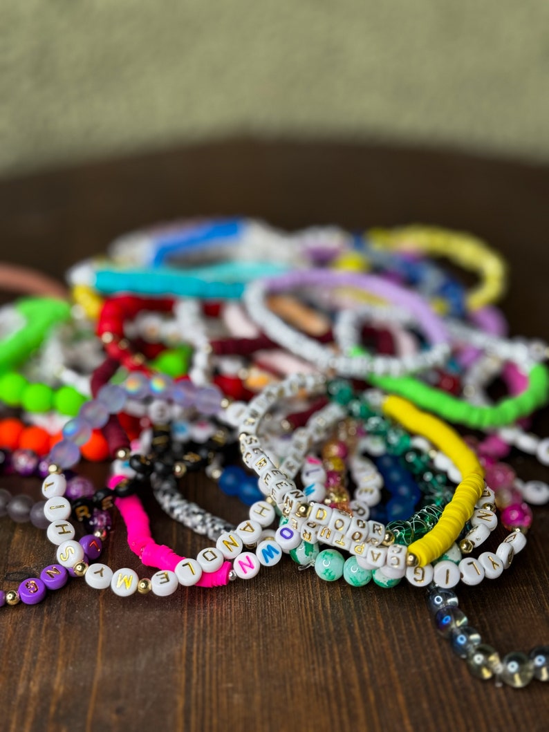 May include: A collection of colorful beaded bracelets with white letter beads spelling out words. The bracelets are in various colors, including pink, yellow, green, and purple. Some bracelets have gold accents and are arranged on a dark wooden surface.