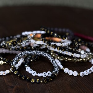 May include: A collection of beaded bracelets with various words and phrases spelled out in black and gold or white and gold letter beads. The bracelets are on a dark wooden surface. Some bracelets have a black and white pattern.