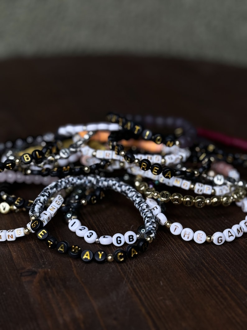 May include: A collection of beaded bracelets with various words and phrases spelled out in black and gold or white and gold letter beads. The bracelets are on a dark wooden surface. Some bracelets have a black and white pattern.