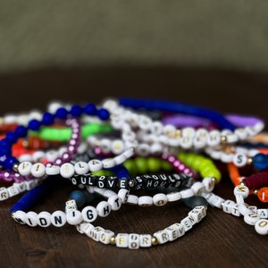 May include: A collection of colorful beaded bracelets with white and black letter beads spelling out words and phrases. The bracelets are in various colors, including blue, green, purple, and orange. The bracelets are on a dark brown wooden surface.