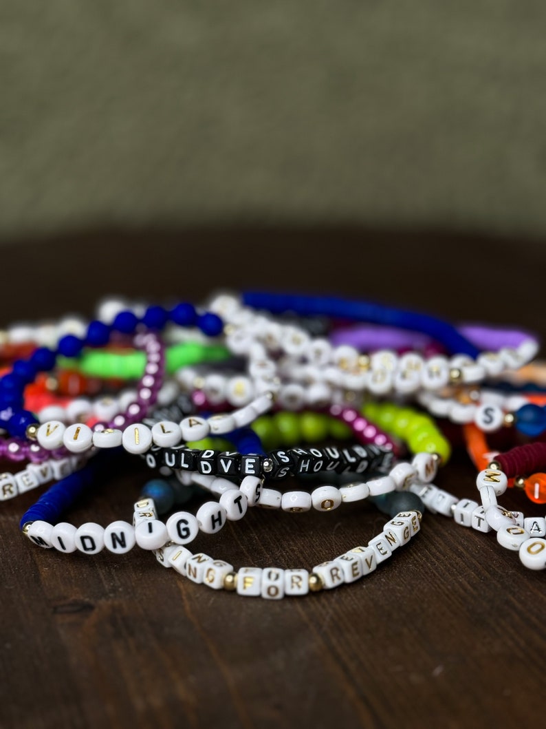 May include: A collection of colorful beaded bracelets with white and black letter beads spelling out words and phrases. The bracelets are in various colors, including blue, green, purple, and orange. The bracelets are on a dark brown wooden surface.