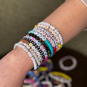May include: A collection of colorful beaded bracelets with white, black, and gold letter beads spelling out phrases like "LOVE STORY" and "CALL IT WHAT YOU WANT." The bracelets are stacked on a wrist, showcasing a variety of colors and styles.