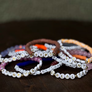 May include: A collection of beaded bracelets with white and gold letter beads spelling out names and words like "TTPD", "ANNIE", and "REALTOR". The bracelets are in various colors, including brown, white, and pink, and are displayed on a dark wooden surface.