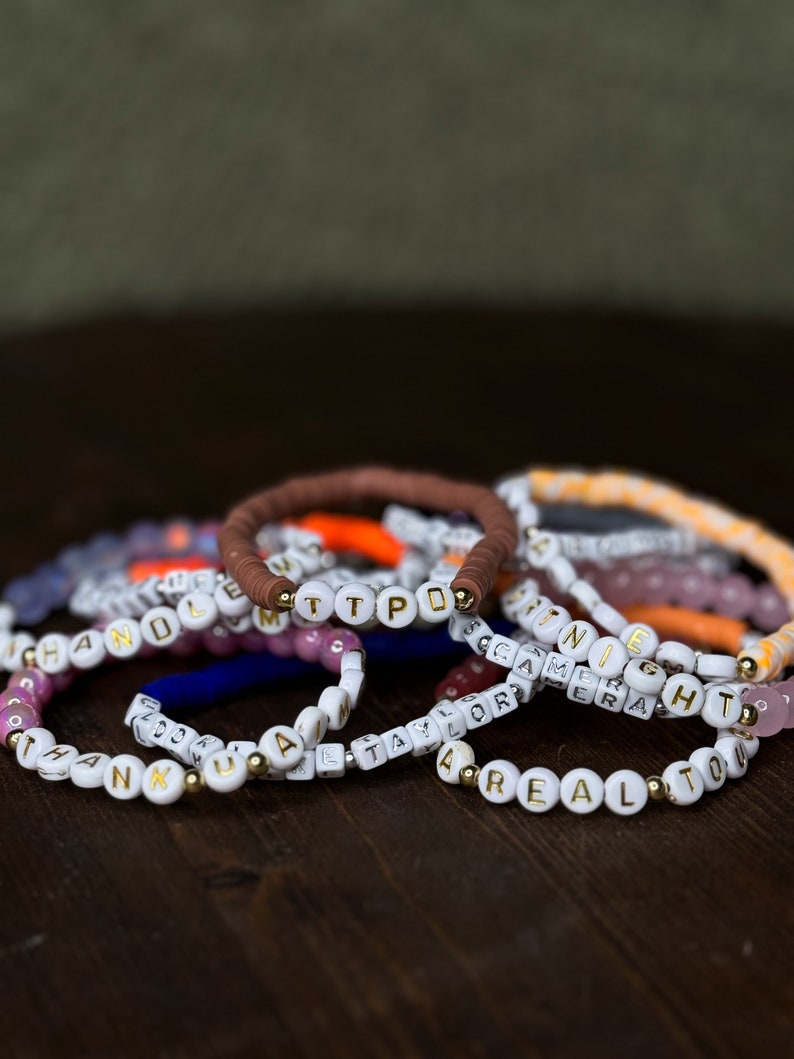 May include: A collection of beaded bracelets with white and gold letter beads spelling out names and words like "TTPD", "ANNIE", and "REALTOR". The bracelets are in various colors, including brown, white, and pink, and are displayed on a dark wooden surface.