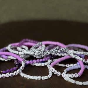 May include: A collection of beaded bracelets in shades of purple, white, and silver. Some bracelets feature letter beads spelling out words like "TIME", "LIVE", and "THE PRETENDER". The bracelets are stacked on a dark wooden surface.