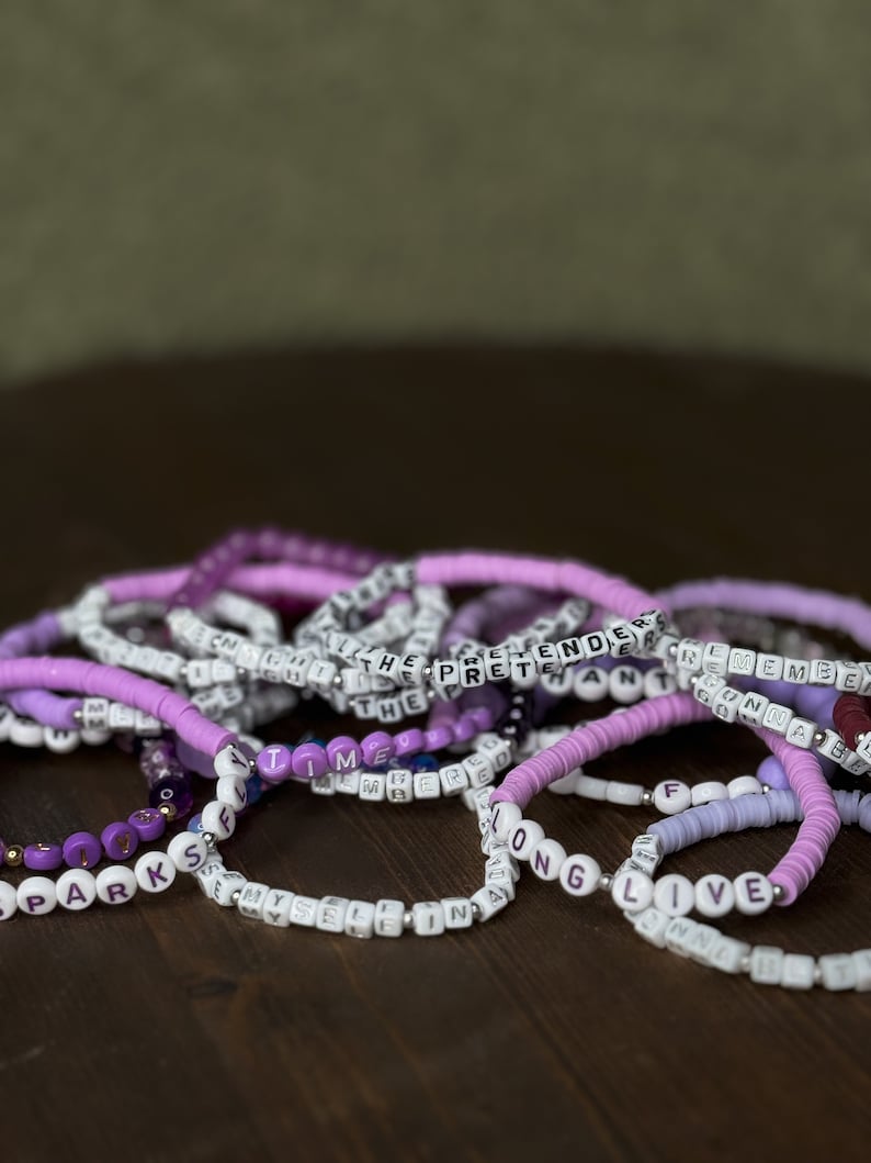 May include: A collection of beaded bracelets in shades of purple, white, and silver. Some bracelets feature letter beads spelling out words like "TIME", "LIVE", and "THE PRETENDER". The bracelets are stacked on a dark wooden surface.