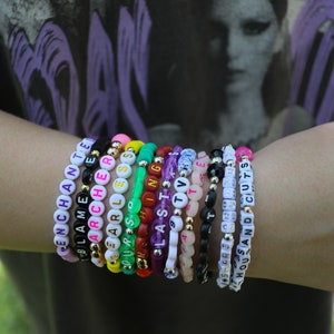 May include: A collection of beaded bracelets with white letter beads spelling out words like "ENCHANTED", "BLAME", "ARCHER", "FEARLESS", "OURS", "LAST", "TV", "STARS", and "HOUSE AND CUTS". The bracelets feature various colors and gold accents.