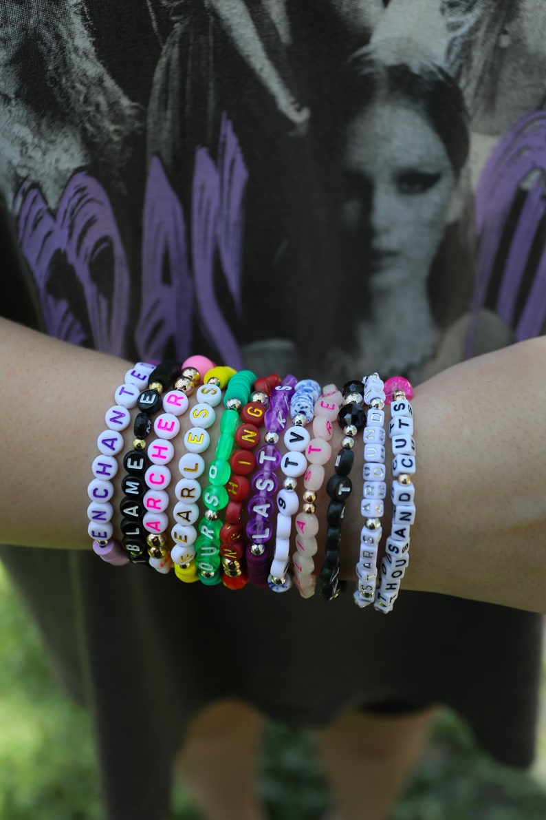 May include: A collection of beaded bracelets with white letter beads spelling out words like "ENCHANTED", "BLAME", "ARCHER", "FEARLESS", "OURS", "LAST", "TV", "STARS", and "HOUSE AND CUTS". The bracelets feature various colors and gold accents.