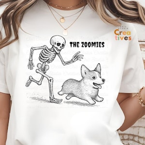 May include: White crew neck t-shirt with a black and white graphic of a skeleton running after a corgi. The text "THE ZOOMIES" is printed above the illustration.