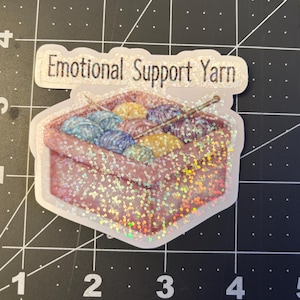 May include: A white sticker with a pink and white glitter background. The sticker features a basket filled with colorful yarn balls and knitting needles. The text "Emotional Support Yarn" is printed on the sticker.