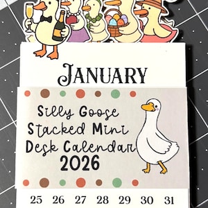 May include: A desk calendar for 2026 featuring cartoon geese. The calendar has the word "January" at the top, with the text "Silly Goose Stacked Mini Desk Calendar 2026" below. The calendar has a white background with colorful dots and a cartoon goose illustration.