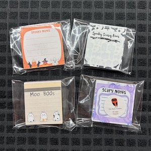 May include: Four individually packaged Halloween-themed sticky note pads. The pads feature various designs, including ghosts, bats, and owls, with phrases like "Spooky Notes" and "Scary Notes". Each pad is in a clear plastic wrapper.