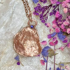 Puede incluir: Un collar con colgante de color cobre con un dije en forma de concha. El dije tiene una superficie texturizada y una pequeña gema redonda de color púrpura. La cadena del collar también es de color cobre. El fondo incluye flores secas en tonos púrpura, rosa y azul.