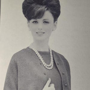 May include: A vintage black and white photograph of a woman wearing a pearl necklace and a matching cardigan and dress. The woman has a bouffant hairstyle and is smiling. The cardigan has buttons.