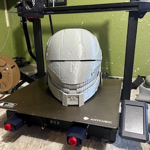 May include: A grey 3D-printed helmet with a detailed, segmented design, on a black 3D printer bed. The printer has a black frame and red and blue accents. The helmet has a face-like structure with a mouth-like opening.