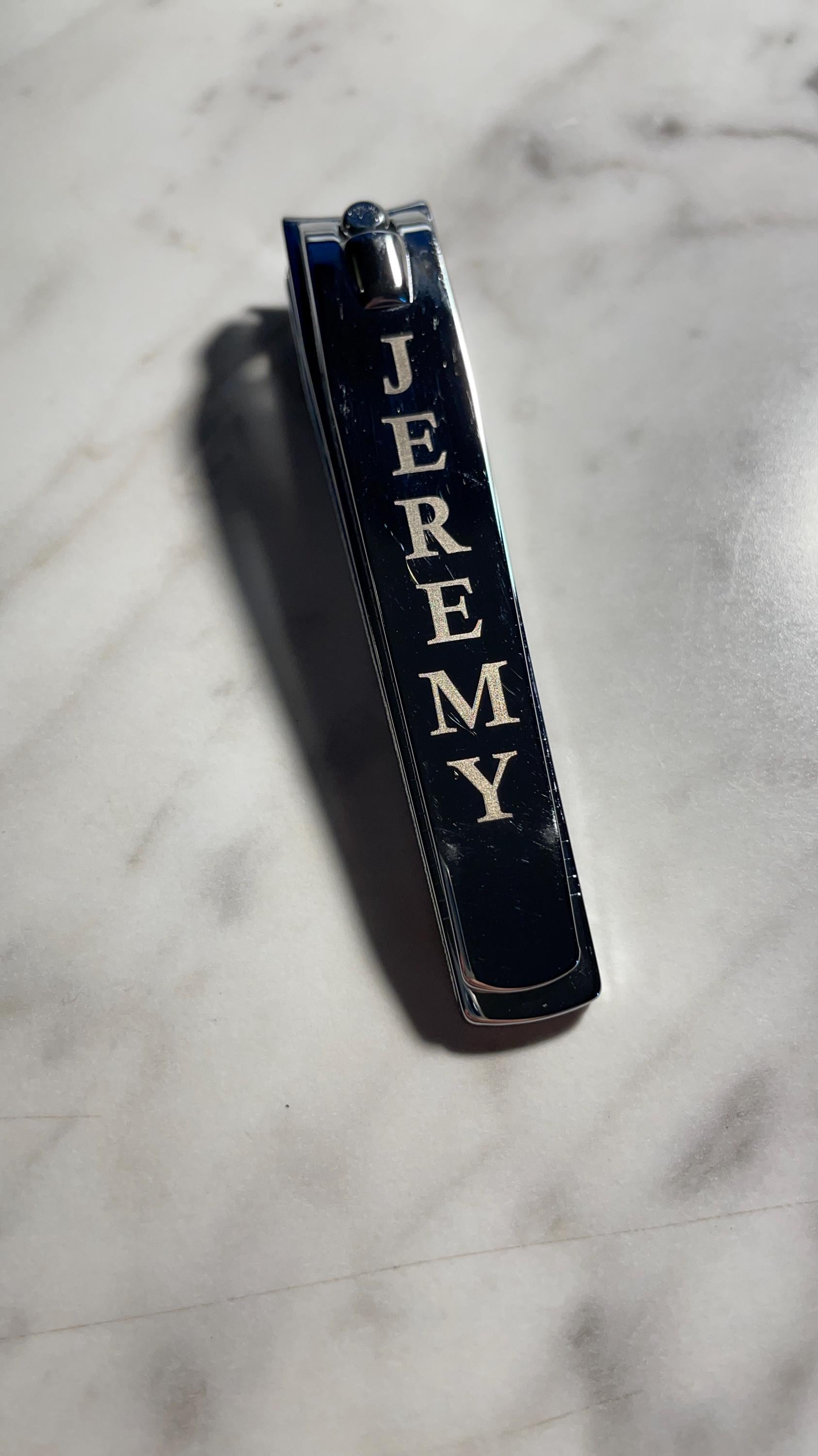 Chrome nail clippers engraved vertically with the name 'JEREMY' on a marble surface.