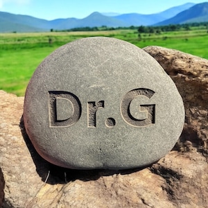 May include: A gray stone with the text "Dr. G" carved into it. The stone is sitting on a rock with a green field and mountains in the background.