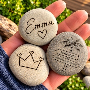 May include: Three smooth, light gray river stones with dark engraved designs. One stone features the name Emma and a heart. Another displays a crown with heart-shaped tips. The third shows a palm tree above two signs reading Punta Gorda and Everything's Better Here.