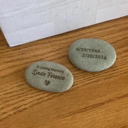 Two engraved river stones on a wooden surface. One says "In Loving Memory Linda Ference" with a heart. The other has dates.