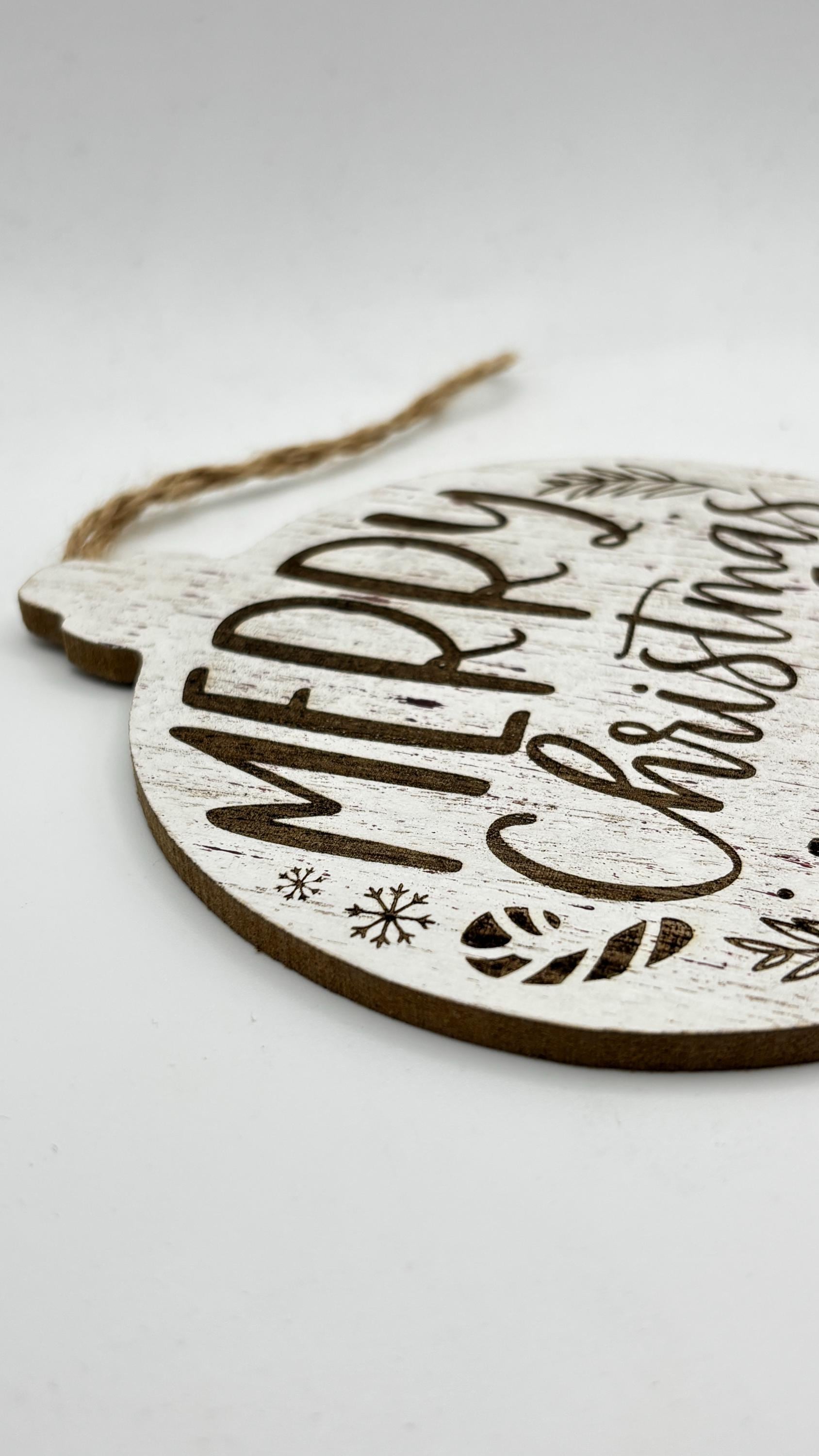 A close-up of a white-washed MDF ornament featuring Merry Christmas engraving with winter motifs.
